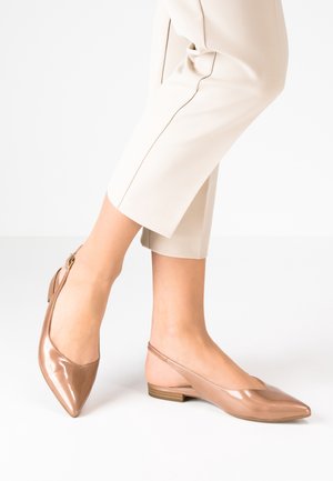 Vrouw draagt beige cropped broek en glanzende nudekleurige lakleren slingback platte schoenen met lage houten hakken.