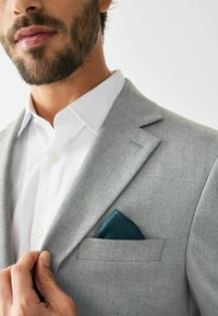 Blazer gris a medida en tejido texturizado, con solapa con muesca, camisa blanca debajo y un pañuelo de bolsillo verde azulado con un patrón geométrico.