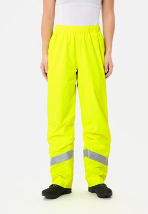 Persoon in felgele hoogzichtbare waterdichte broek met reflecterende banden boven zwarte schoenen, staande tegen een effen witte achtergrond.