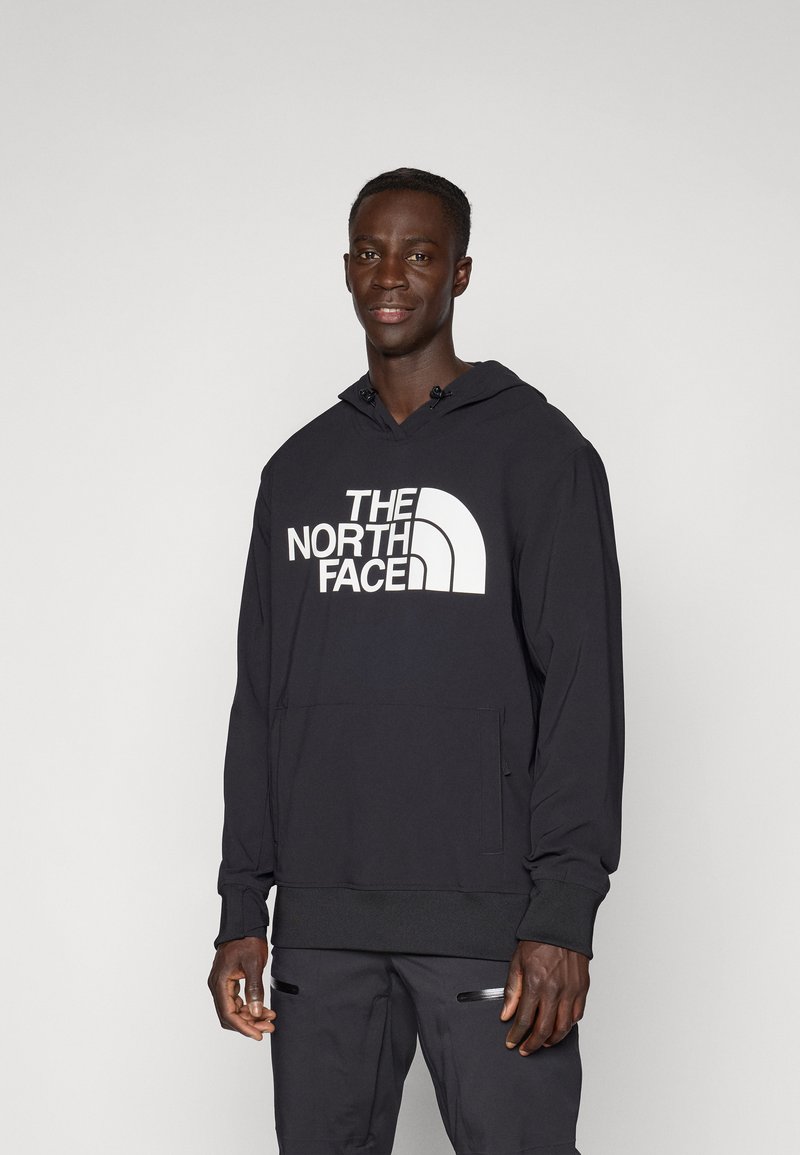 Zwarte hoodie van zacht materiaal, met een kangoeroezak, ribgebreide manchetten en zoom, en een groot wit logo op de voorkant.