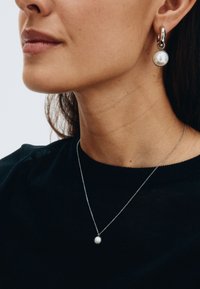 Orecchini a cerchio in argento con dettagli in perle bianche; una delicata collana in argento con un piccolo pendente a perla bianca. Maglione nero visibile.