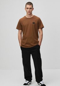 Brązowa bawełniana koszulka z krótkim rękawem, z małą czarną metką na piersi. Połączona z czarnymi spodniami cargo i czarno-białymi trampkami.