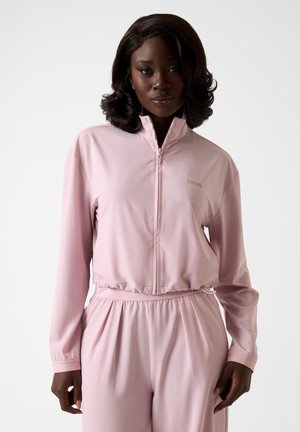 Donna con capelli ricci fino alle spalle che indossa una giacca rosa chiaro con zip e pantaloni abbinati, su uno sfondo semplice.