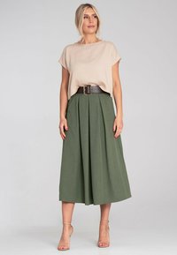 Beżowa bluzka z krótkim rękawem o luźnym kroju, zestawiona z zieloną plisowaną spódnicą midi oraz szerokim czarnym paskiem z metalową klamrą w kolorze srebrnym. Przejrzyste, neutralne tło.