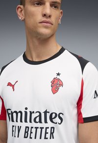 Hvid kortærmet trøje med sorte og røde detaljer, Puma-logo, et drag emblemet og teksten "Emirates Fly Better". Glat tekstur.