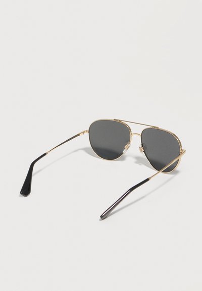 Ένα ζευγάρι γυαλιά ηλίου aviator με χρυσό πλαίσιο, μαύρους βραχίονες και σκουρόχρωνους γκρι φακούς σε λευκό φόντο.