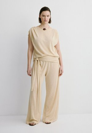 Mujer con top sin mangas beige con lazo lateral y pantalones de pierna ancha, combinados con un único collar oscuro llamativo y sandalias.