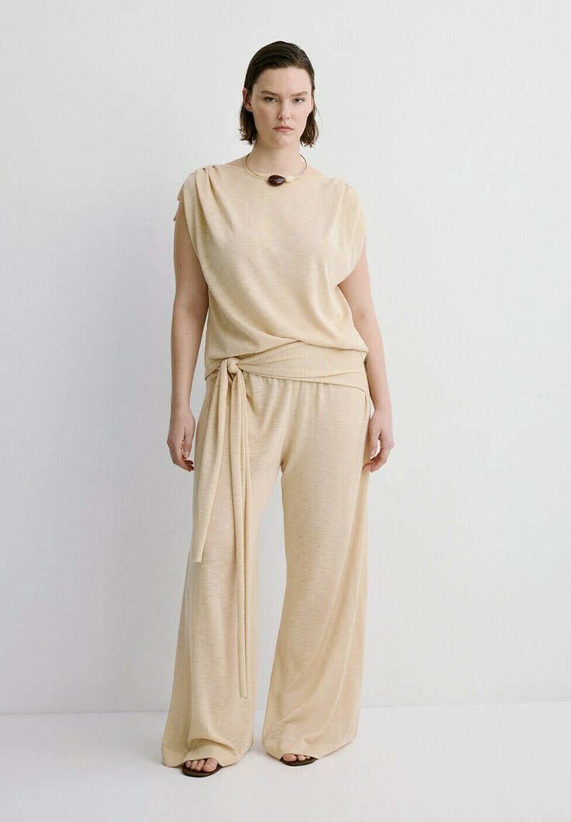 Femme portant un haut beige sans manches avec nouage sur le côté et un pantalon à jambes larges, assortis d'un unique collier sombre et de sandales.