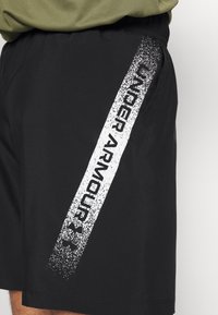Mustat urheilusortsit, joissa on teksturoitu valkoinen Under Armour -logo painettuna pystysuoraan sivuun, ja ne ovat valmistettu sileästä, kevyestä kankaan. 