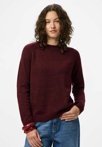 Burgundski pleteni pulover z okroglo izrezom in dolgimi rokavi, v kombinaciji z modrimi kavbojkami. Pulover ima mehak, teksturiran videz.