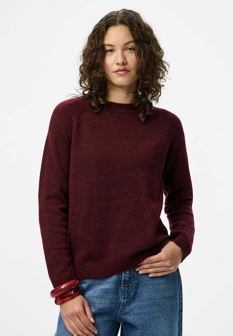 Burgundski pleteni pulover z okroglo izrezom in dolgimi rokavi, v kombinaciji z modrimi kavbojkami. Pulover ima mehak, teksturiran videz.