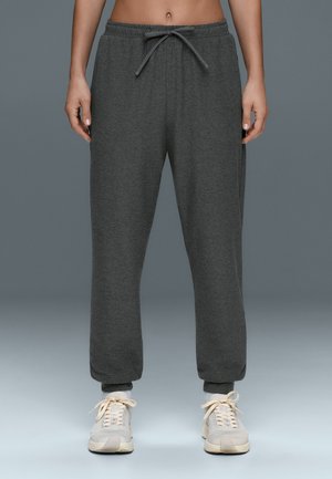 Persoon draagt donkergrijze sweatpants met aantrekkoord in de taille en lichtbeige sneakers, staand tegen een effen grijze achtergrond.