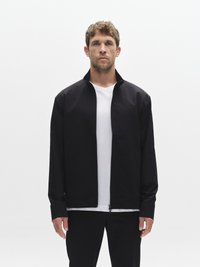 Melnā rāvējslēdzēja jaka no gludas auduma ar augstu apkakli un gariem piedurknēm, komplektā ar baltu T-kreklu un melnām biksēm.