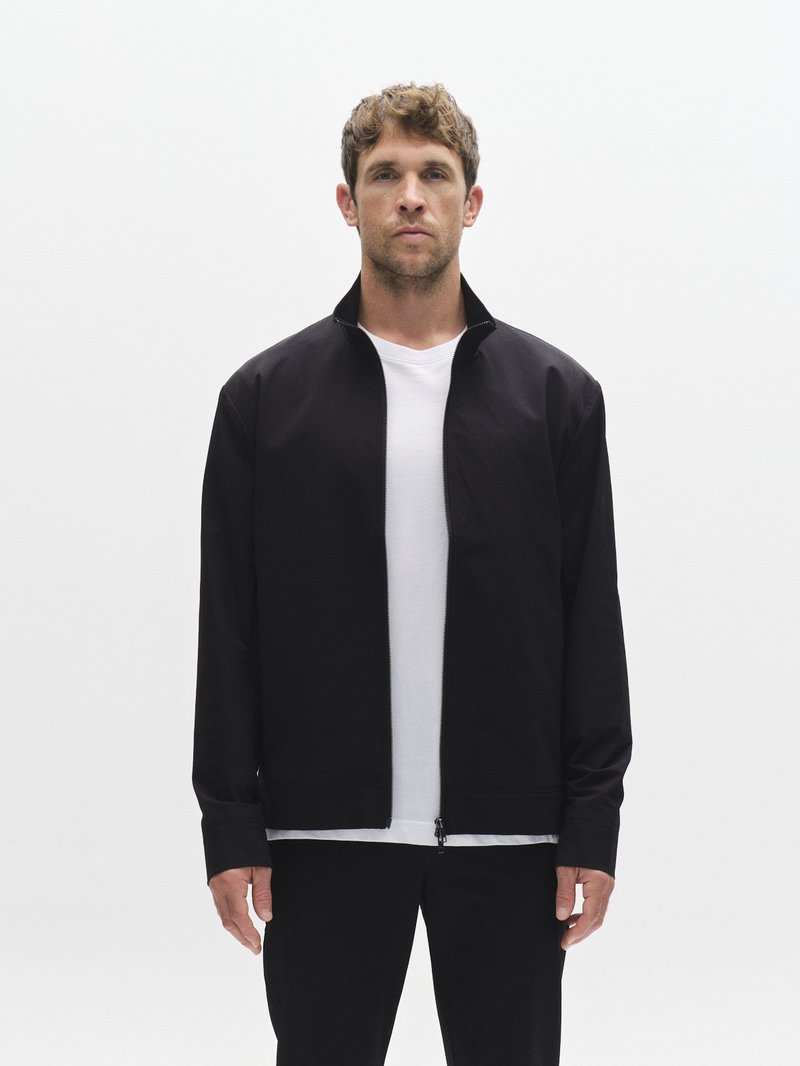 Melnā rāvējslēdzēja jaka no gludas auduma ar augstu apkakli un gariem piedurknēm, komplektā ar baltu T-kreklu un melnām biksēm.