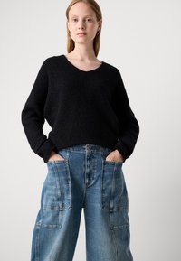 Suéter negro texturizado con escote en V, combinado con amplios pantalones de mezclilla azul claro que presentan grandes bolsillos frontales y detalles de costura visibles.