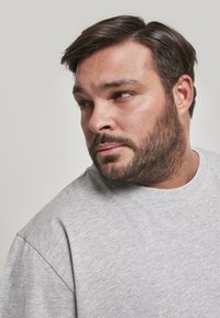 T-shirt grise en laine avec un col rond et des manches courtes, fabriquée en tissu doux et texturé. Modèle avec des cheveux foncés et une barbe, vu de profil.