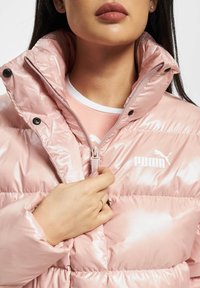 Rožinė pūkinė striukė su blizgiu paviršiumi, turinti aukštą apykaklę, užtrauktuką ir juodus spaudimo mygtukus. Logotipas ant krūtinės. Minkšta audinio tekstūra.