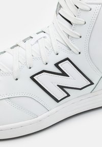 Weiße hoch geschnittene Sneakers aus Leder, mit schwarzen Akzenten, einem großen weißen "N"-Logo, strukturierten Ösen und flachen weißen Schnürsenkeln.