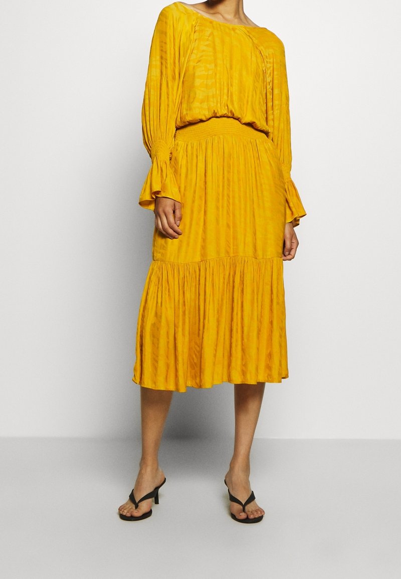 Yellow dress made of lightweight, textured fabric; features long sleeves, a fitted waist, and a tiered, flared skirt. Worn with black sandals.