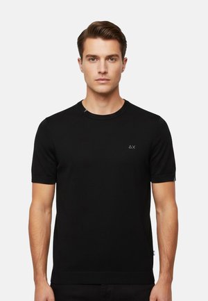 Giovane uomo di fronte che indossa una maglietta nera a maniche corte, semplice, con un piccolo logo bianco a forma di triangolo e X sul petto.