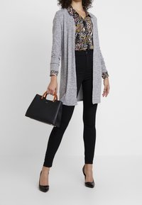 Un long cardigan gris sur un chemisier imprimé, un jean skinny noir et des talons aiguilles noirs. Un sac à main noir avec des poignées beiges, au design épuré.