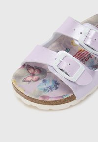 Λιλά σανδάλι με δύο ρυθμιζόμενες λευκές αγκράφες και εσωτερική σόλα με σχέδιο λουλουδιών και πεταλούδες σε φελλό σόλα.