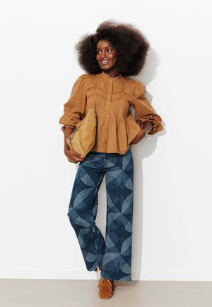 Femme aux cheveux bouclés portant une blouse marron boutonnée, un jean large à motifs, des mocassins marron, et tenant un sac en daim beige contre un mur blanc.