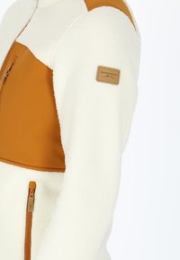 Fleece jacket with a cream body and brown upper panel. Features a zippered pocket and a logo patch on the sleeve. Soft texture.