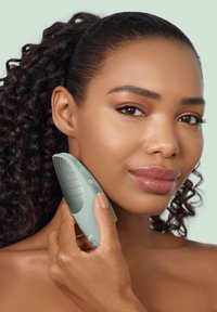 Dispositivo per la pulizia del viso di colore verde tenue, forma ergonomica, superficie testurizzata per una presa sicura, tenuto contro la guancia, con il tono di pelle naturale del modello visibile.