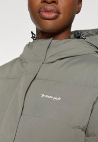 Pilkas pūkinis švarkas su aukštu apykakle, ant krūtinės siuvinėtu "snow peak" logotipu ir raišteliais pritaikytiems reguliuoti.