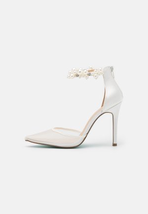 Bílé saténové lodičky na vysokém podpatku s špičatou špičkou, které mají síťovanou část a kotníkový pásek zdobený perlami a dekorativními prvky.