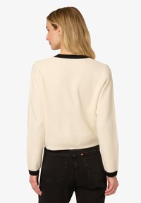 Cremfarbener Pullover mit strukturiertem Finish, ausgestattet mit schwarzen Rippbündchen am Hals und an den Ärmeln. Kombiniert mit dunklen Jeans, die eine lässige Passform zeigen.