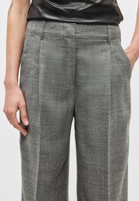 Calças cinzentas feitas à medida com tecido texturizado. Apresentam pregas frontais e bolsos laterais, desenhadas para um ajuste clássico.