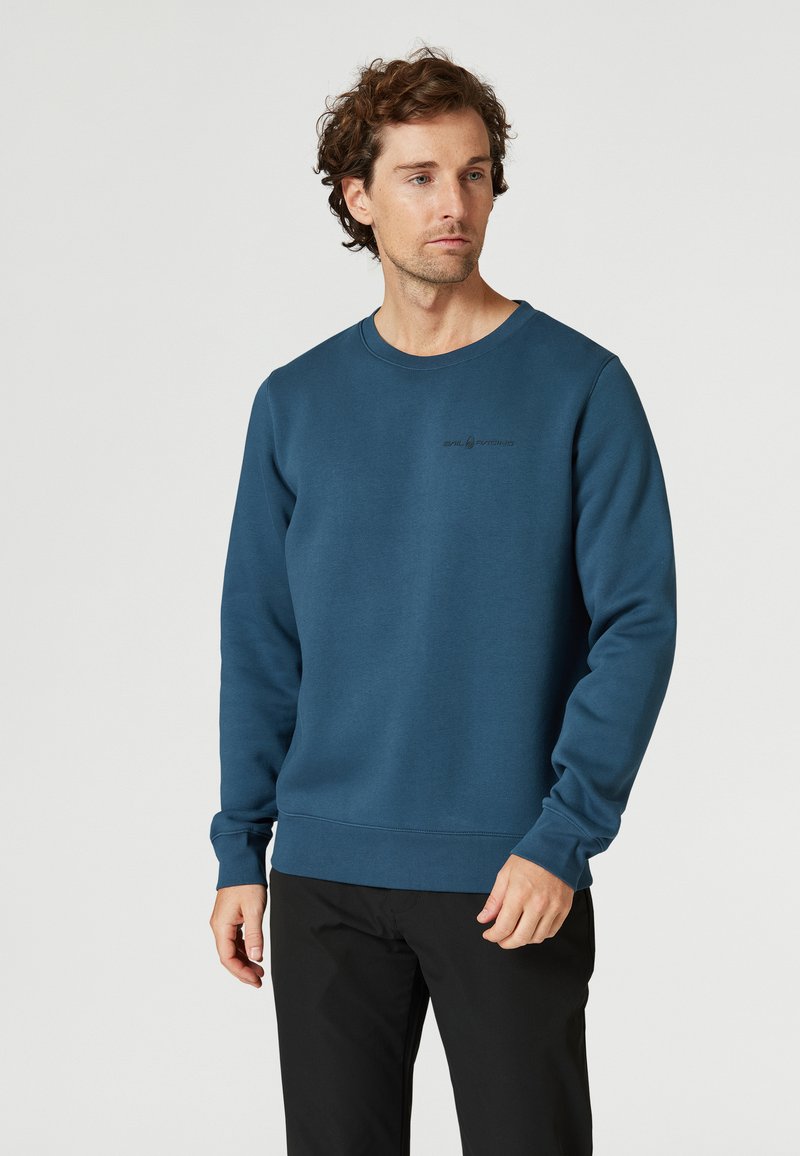 Sudadera azul hecha de una tela suave, con cuello redondo y puños de canalé. Un pequeño logo es visible en el pecho. Se combina con pantalones negros.