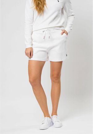 Kvinde iført hvid sweatshirt, hvide shorts med snører og hvide sneakers, står med den ene hånd i lommen mod en lys baggrund.