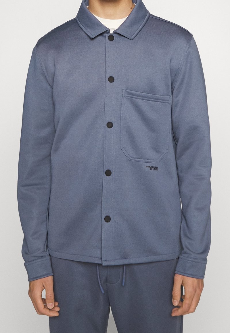 Homme portant une veste bleue boutonnée avec une poche poitrine et un pantalon bleu assorti sur un fond uni.