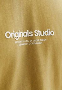 Khaki bomuldsstof med hvid tekst, der skriver "Originals Studio", med yderligere tekst til brand- og designoplysninger nedenfor.