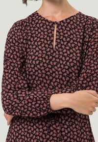 Dunkel gemustertes Kleid mit einem Paisleymuster in Rot und Creme. Verfügt über eine Schlüsselöffnung auf der Vorderseite und lange, puffige Ärmel. Hergestellt aus weichem Stoff.