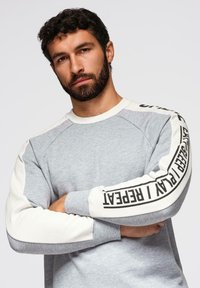 Sudadera gris con mangas raglán blancas, con un destacado patrón de texto en negro en el brazo izquierdo, hecha de una tela suave.
