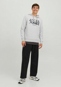 Sudadera gris con un estampado de logotipo, capucha con cordón, puños acanalados, llevada sobre una camiseta oscura, combinada con pantalones anchos negros y zapatillas.