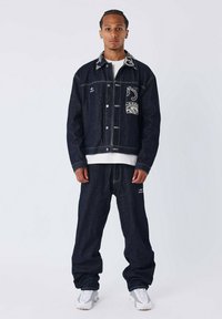 Man in een donkerdenimjack met een patroon op de kraag en zak, bijpassende losse jeans, witte sneakers, staand tegen een effen achtergrond.