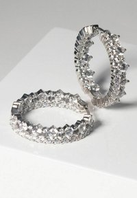 Silverörhängen i ringform med flera klara ädelstenar och en design med vågiga kanter. Glittrande yta och cirkulär form.