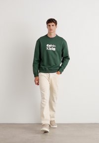 Felpa verde con logo "Calvin Klein", jeans color crema e sneakers beige. Design semplice con maniche lunghe e scollo rotondo.