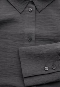 Camicia button-up grigio scuro in tessuto liscio e strutturato. Presenta un colletto a punta e due bottoni ai polsini. Design leggermente spiegazzato.