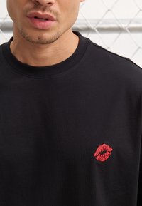 Svart bomull T-skjorte med rund hals og korte ermer, med et lite rødt brodert leppestift-kyssemotiv på venstre side.