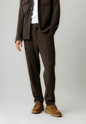 Hombre con pantalones de punto marrón oscuro, chaqueta de camisa abotonada a juego, camisa blanca y zapatos de ante marrón claro con cordones, de pie con una mano en el bolsillo.