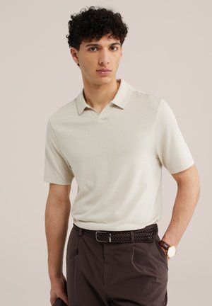 Mann mit lockigem Haar, der ein beiges kurzärmeliges Poloshirt, braune Nadelstreifenhose, geflochtenen Gürtel und Armbanduhr trägt, steht mit einer Hand in der Tasche.