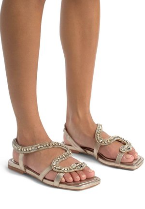 Sandali piatti beige con una finitura lucida e testurizzata. Elegante cinturino ondulato decorato con embellishments trasparenti e design a punta quadrata.