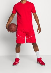 Hombre en ropa deportiva roja sosteniendo un balón de baloncesto con las piernas separadas, vistiendo calcetines blancos y zapatillas de baloncesto rojas frente a un fondo liso.