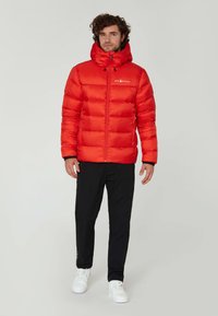 Rode gewatteerde jas met capuchon, voorzien van een ritssluiting aan de voorkant en elastische manchetten. Wordt gedragen over zwarte broek en witte sneakers.
